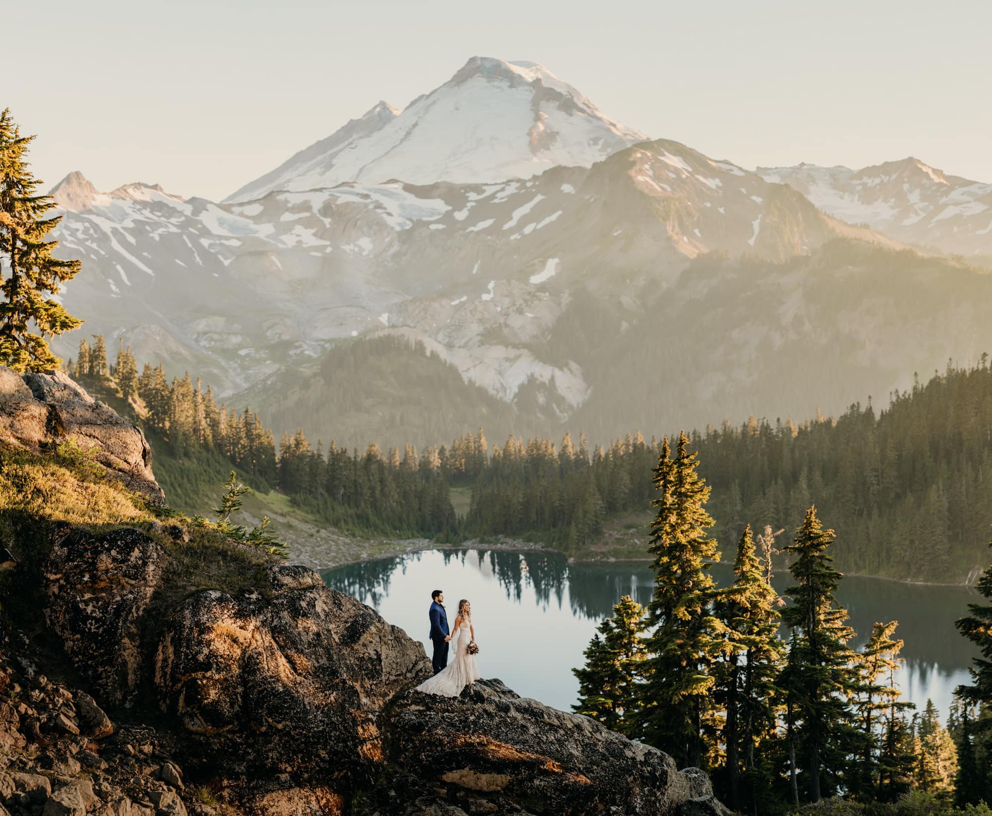 A couple stands together in the mountains overlooking a lake.