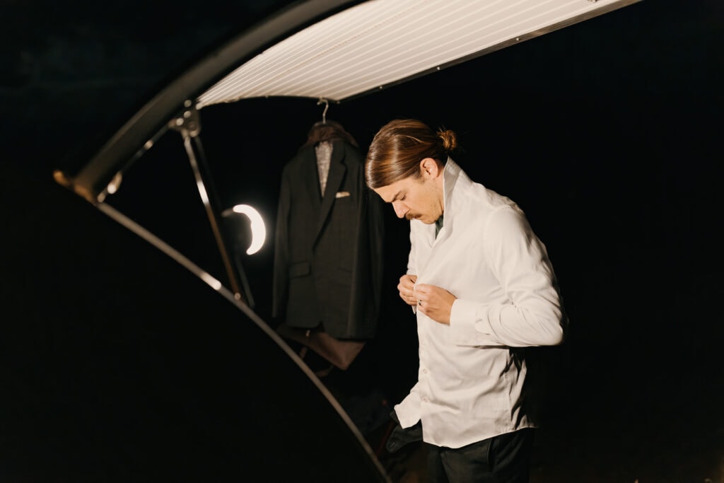 A detail photo of the groom buttoning his shirt.