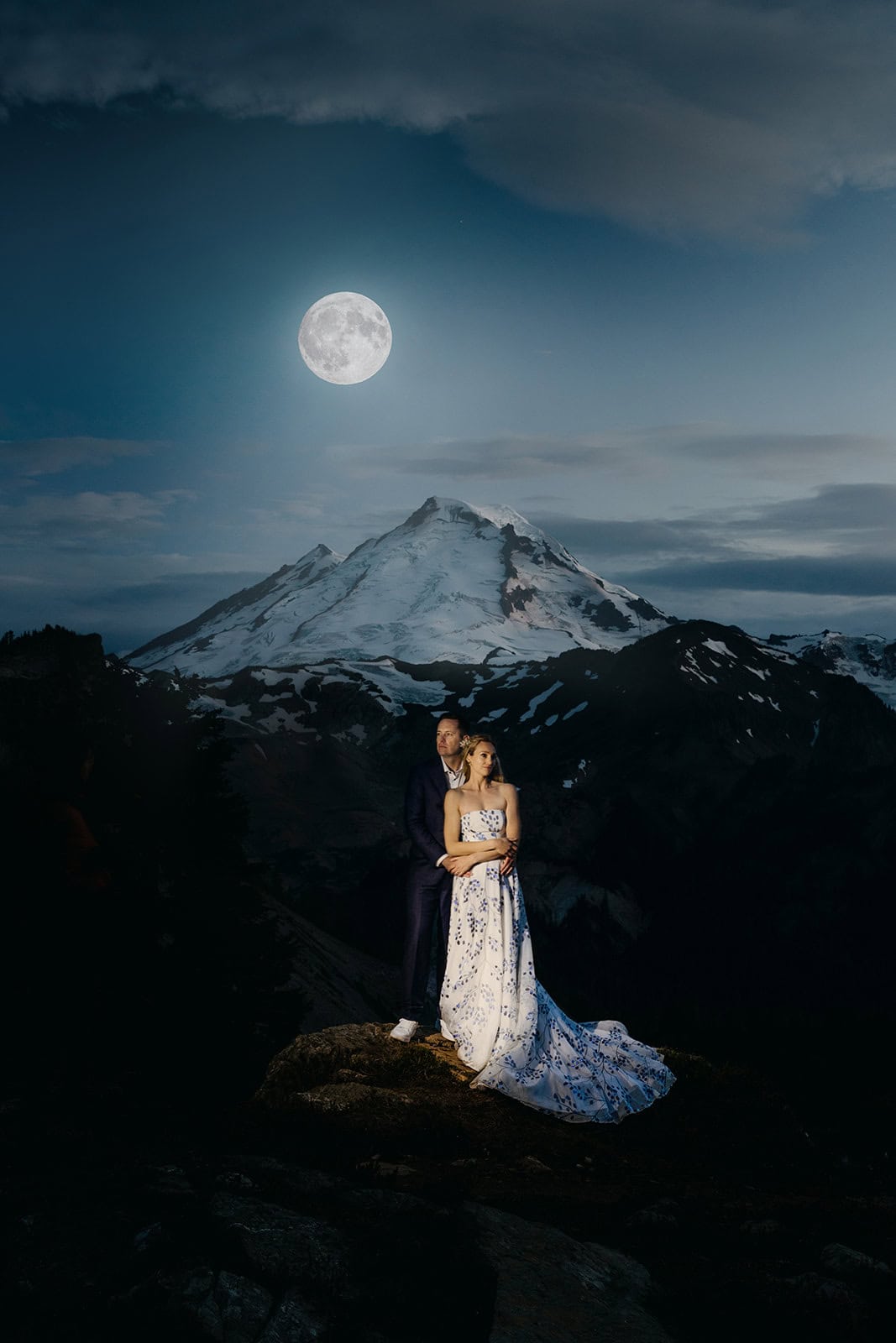 A couple stands under the moonlight in the mountains together on their wedding night.