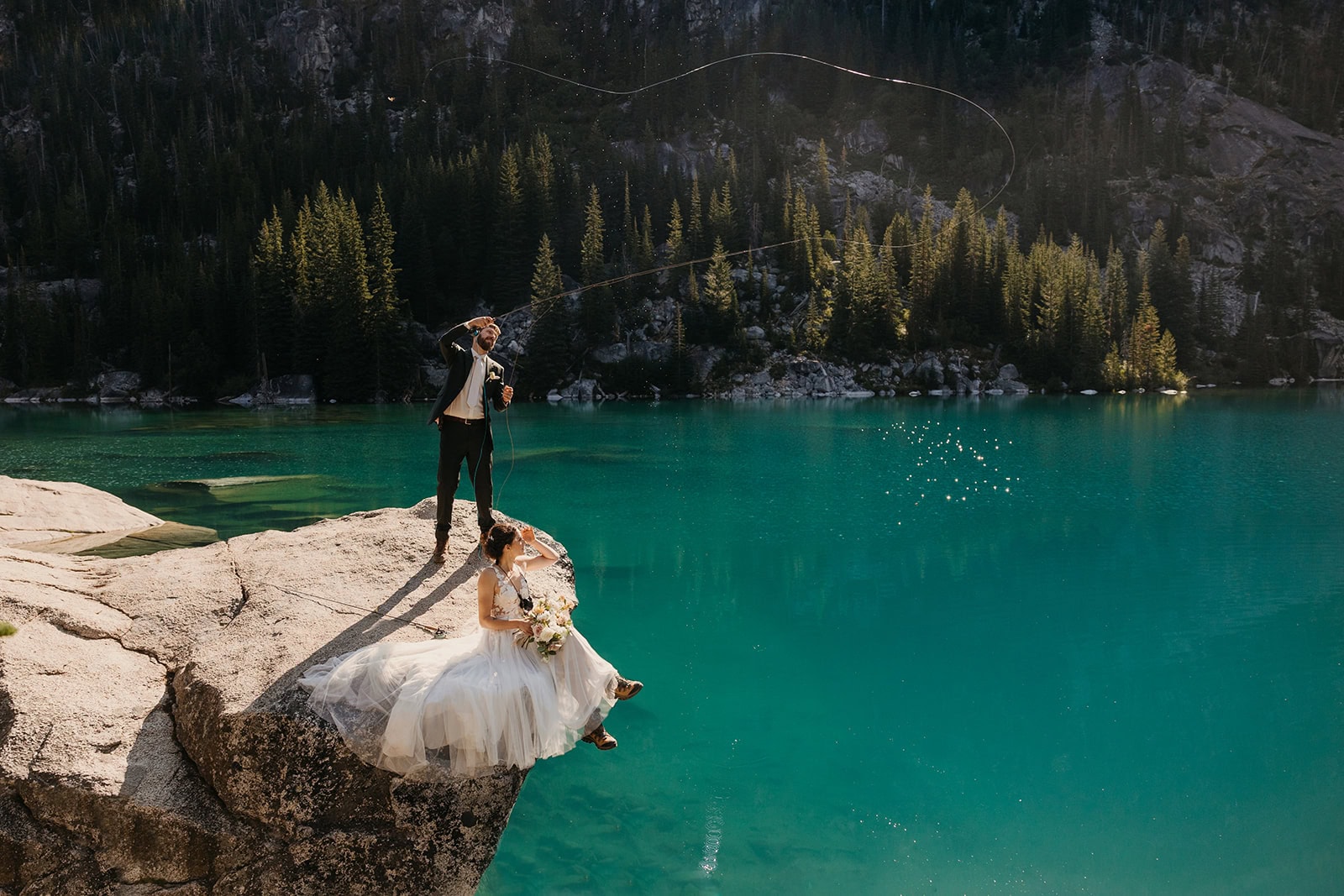 A bride and groom together on a rock atop a turquoise colored lake where the groom is fly fishing.