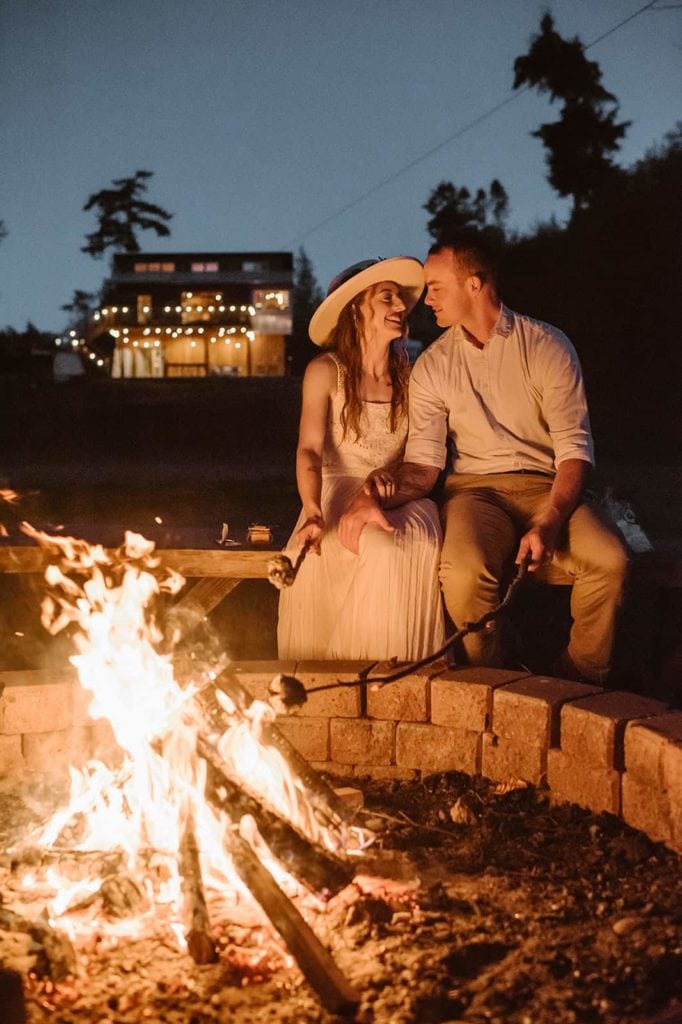 A couple makes smores by a camp fires with a cabin in the background.