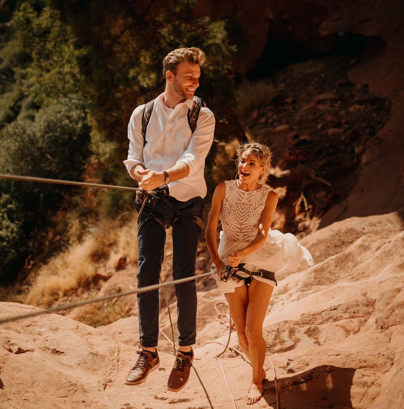 A newlywed couple walking up the side of a rock while holding onto ropes
