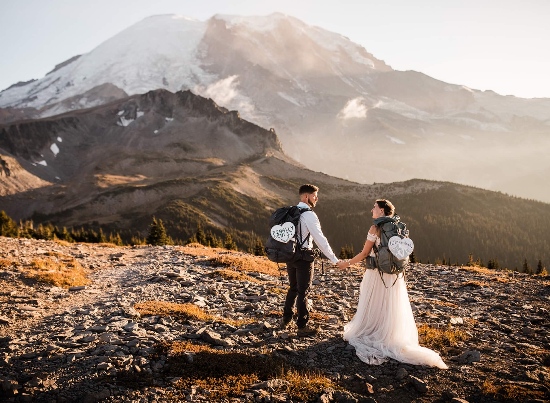 A newlywed couple holding hands in the mountains with hiking backpacks on 