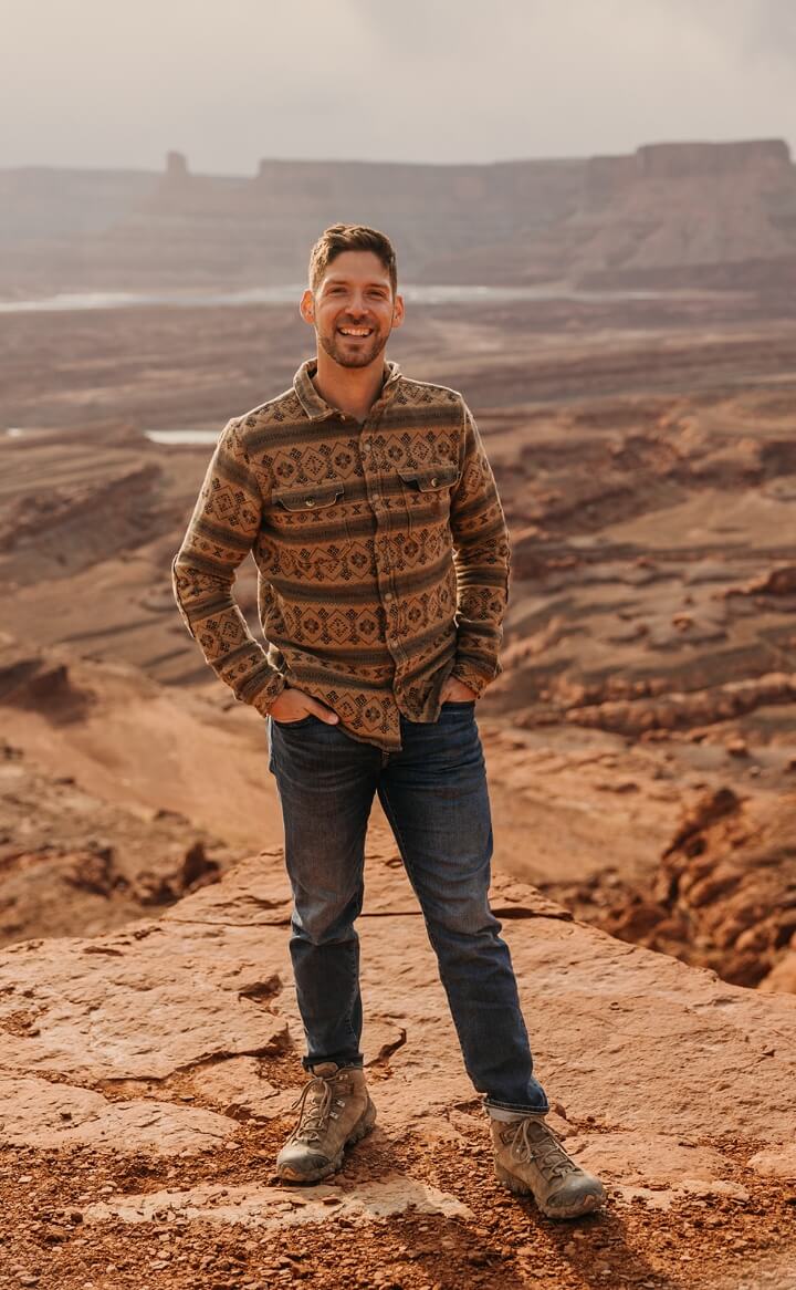 A person smiling with their hands in their pockets while standing at the top of a rocky canyon 