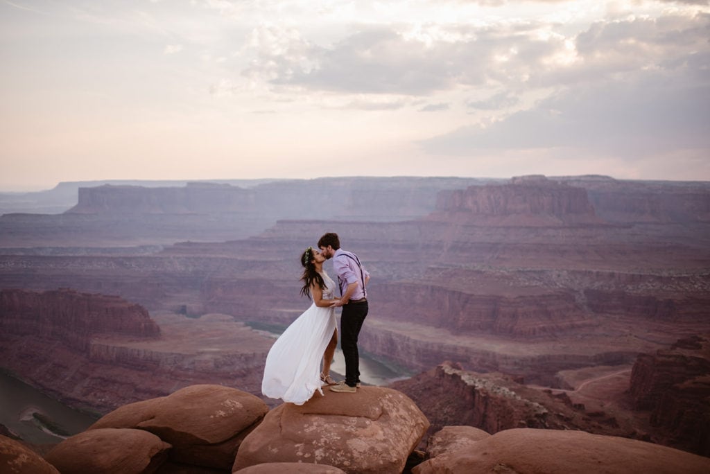 A bride and groom kiss as the wind catches the bride's dress.