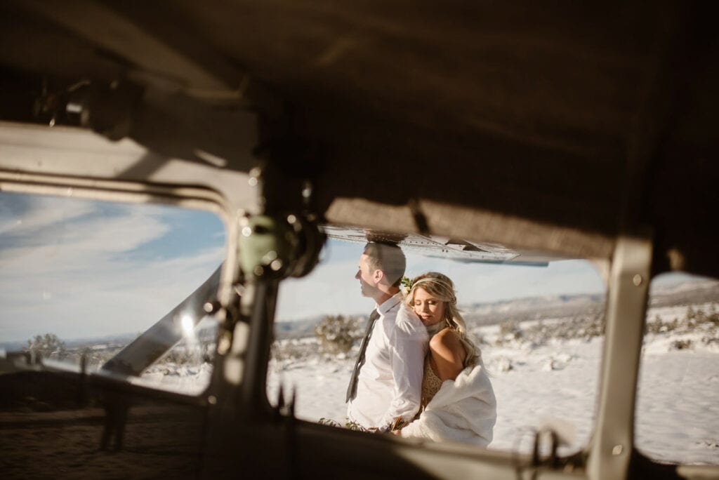 A bride hugs her groom as they stand in the snow on a sunny day outside of the plan that chartered them. 