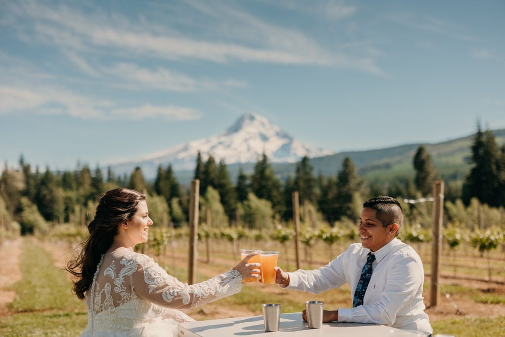 Two brides cheers their beer together on their wedding day at a winery with mountain views.