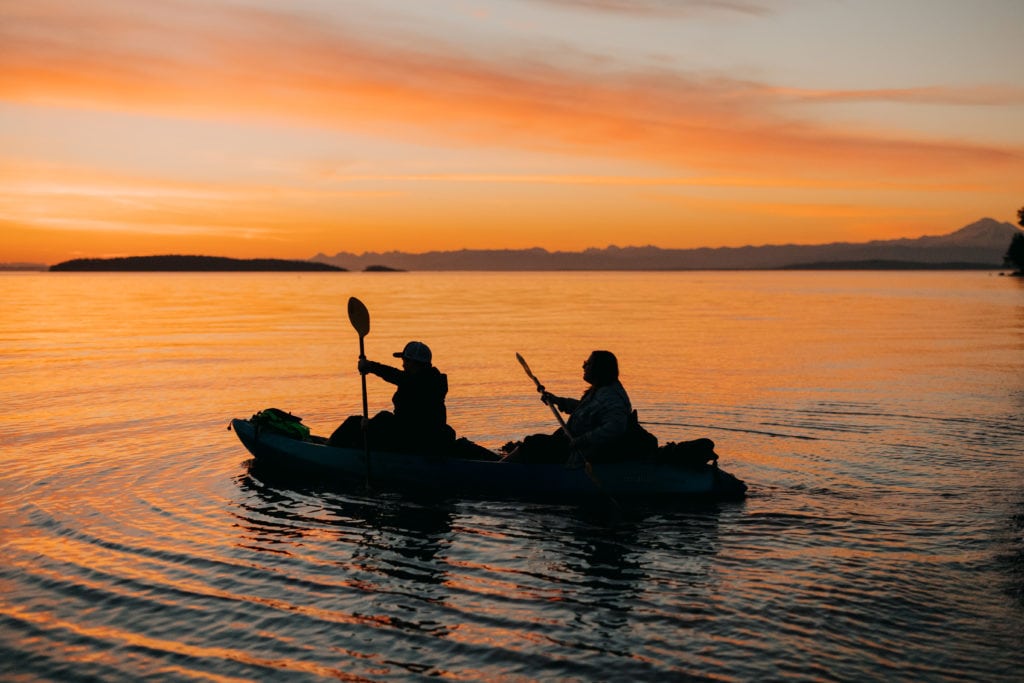 the bride and groom kayak, sending ripples around, in the orange sunset.