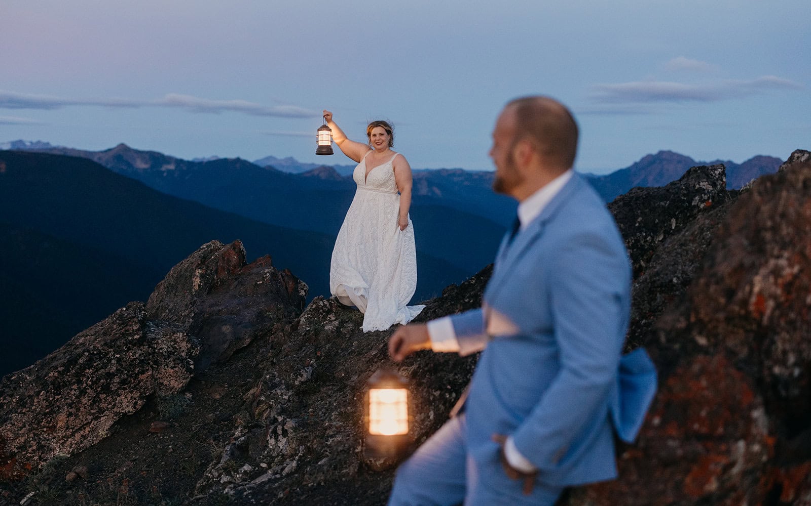 A bride holds a lantern and looks toward her groom as they watch sunrise in the mountains.