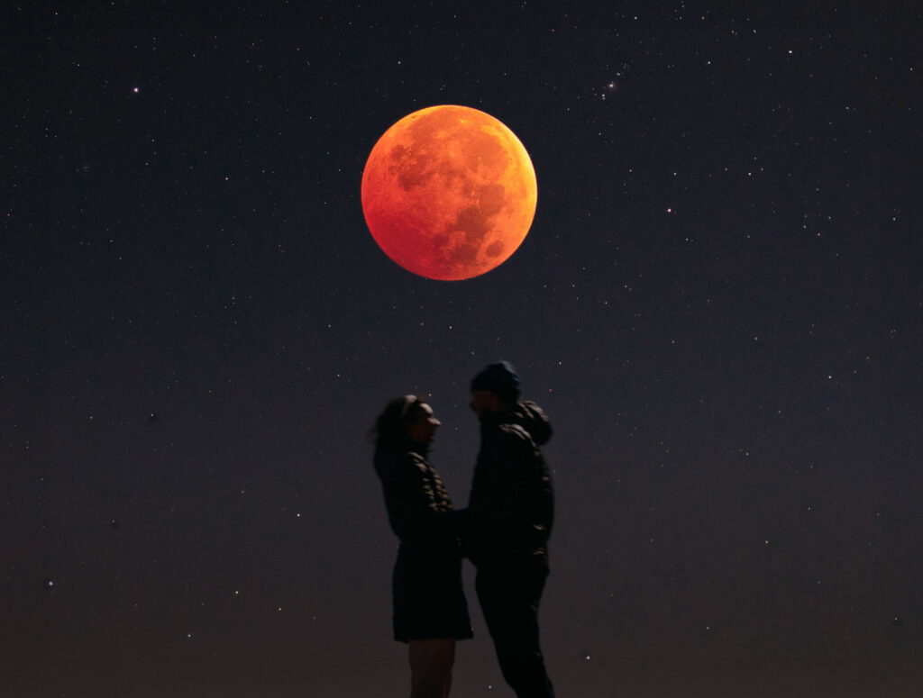 A couple stands together with the full moon above them high in the sky.