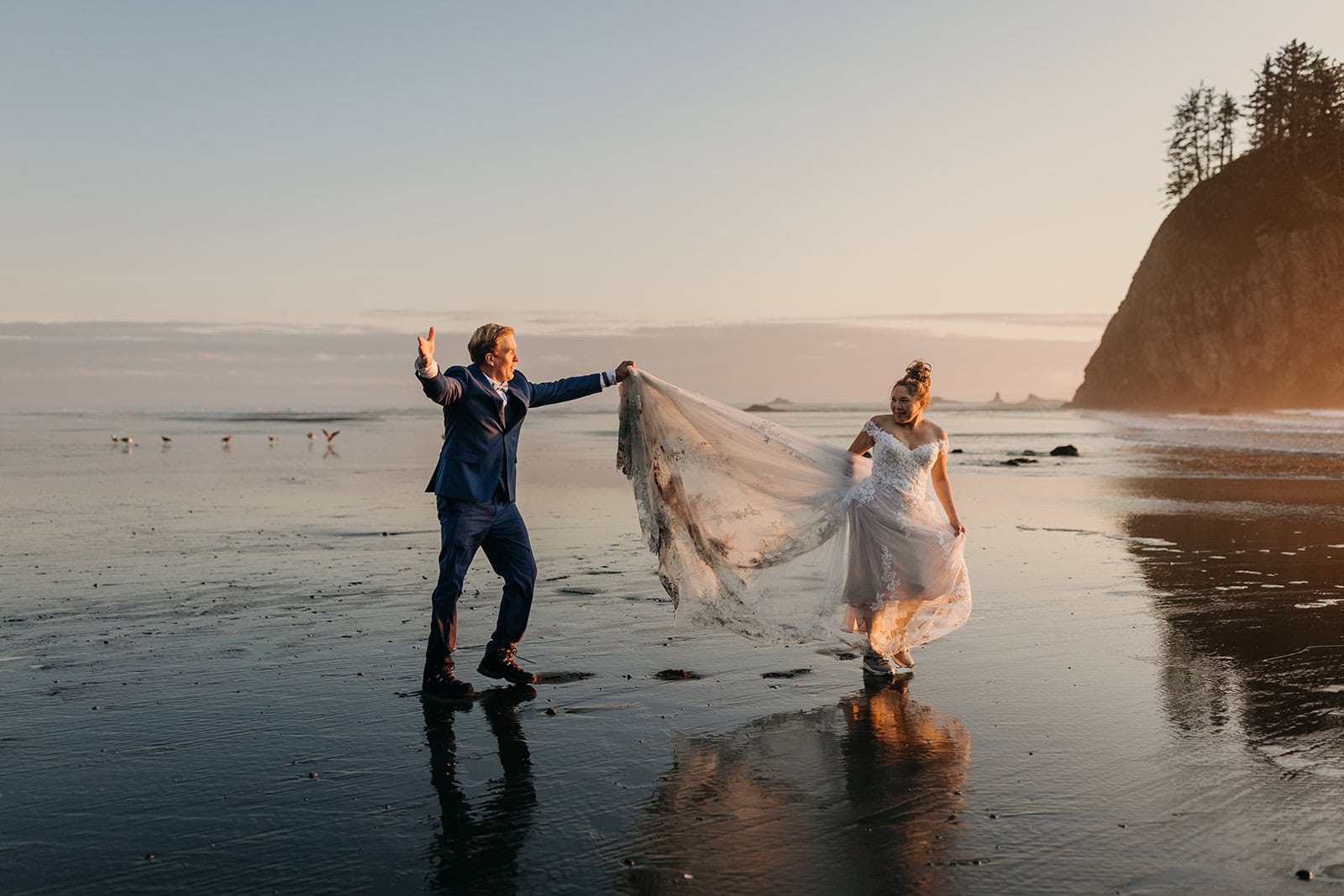 A groom carries a brides train of her dress as they walk across the beach.