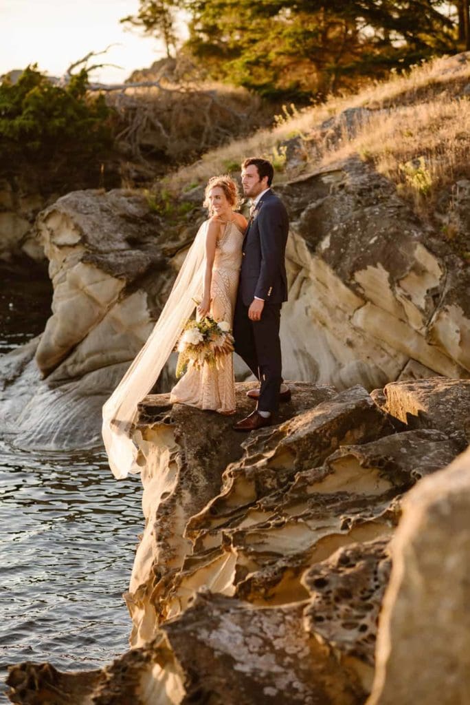 A bride and groom stand together on a ledge right off the ocean shore.