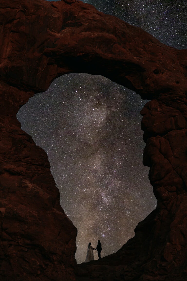 A bride and groom stand together at Arches National Park with the Milkyway passing through Turret Arch behind them
