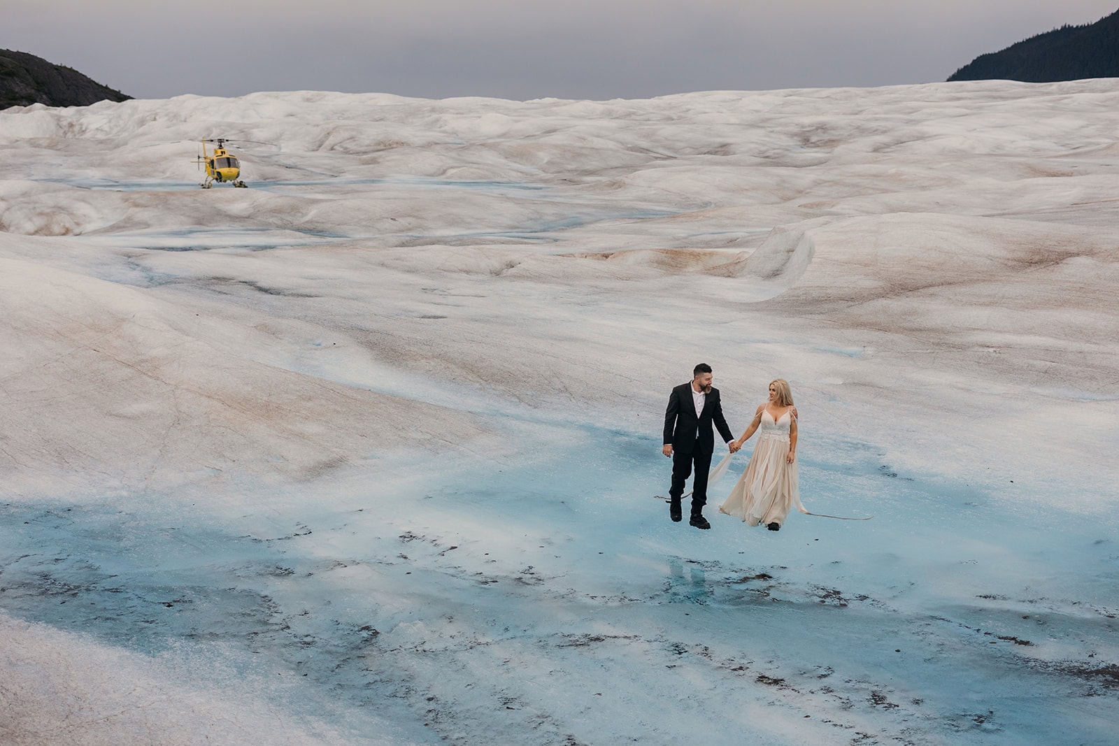 A couple walks across a glacier that they arrived at via a yellow helicopter in the background.