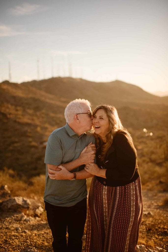 A groom kisses his bride on the temple as they stand on a sand cliff outlook over the canyons in Moab. 