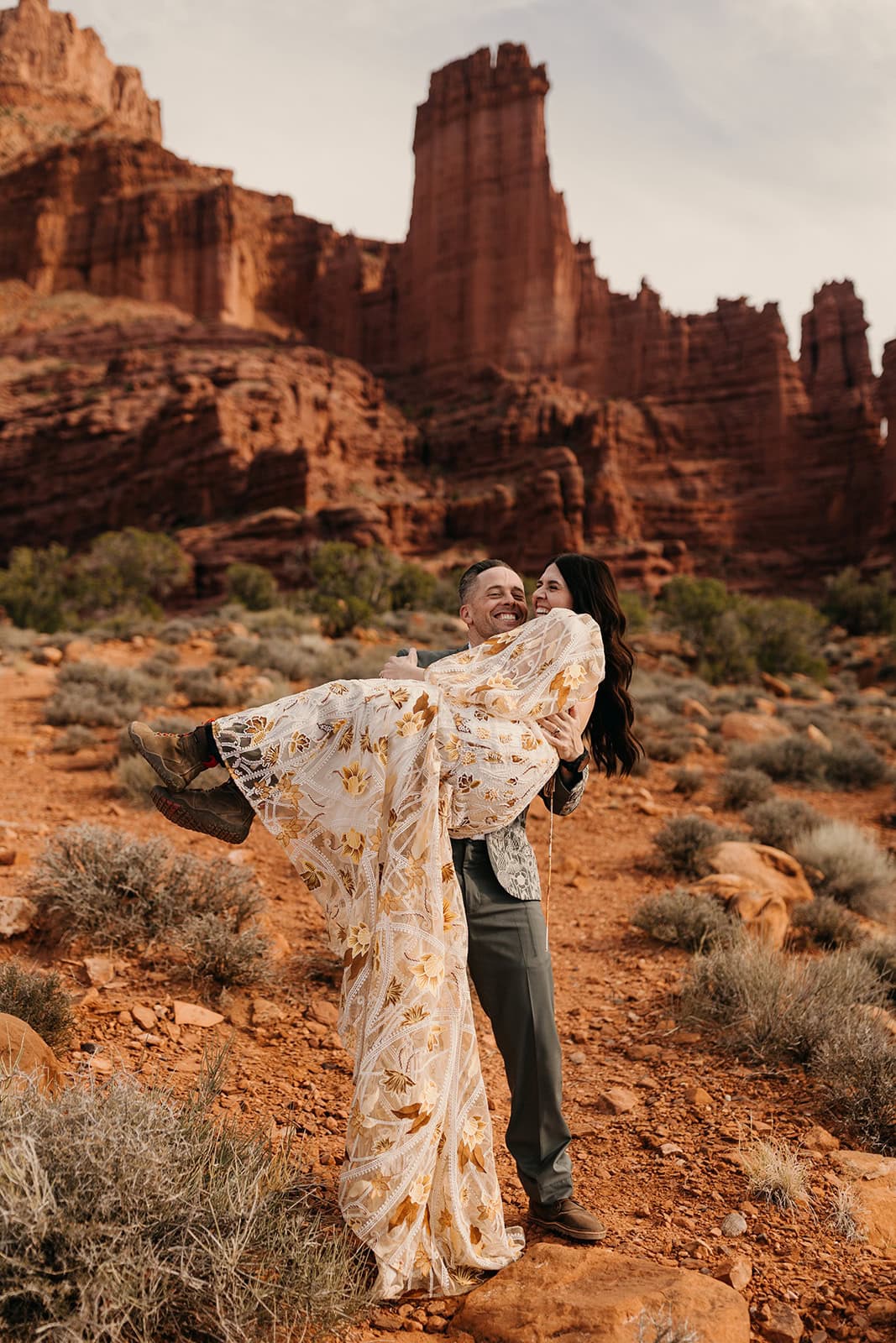 A groom carries his bride down the trail smiling, her Rue De Seine dress making a statement.