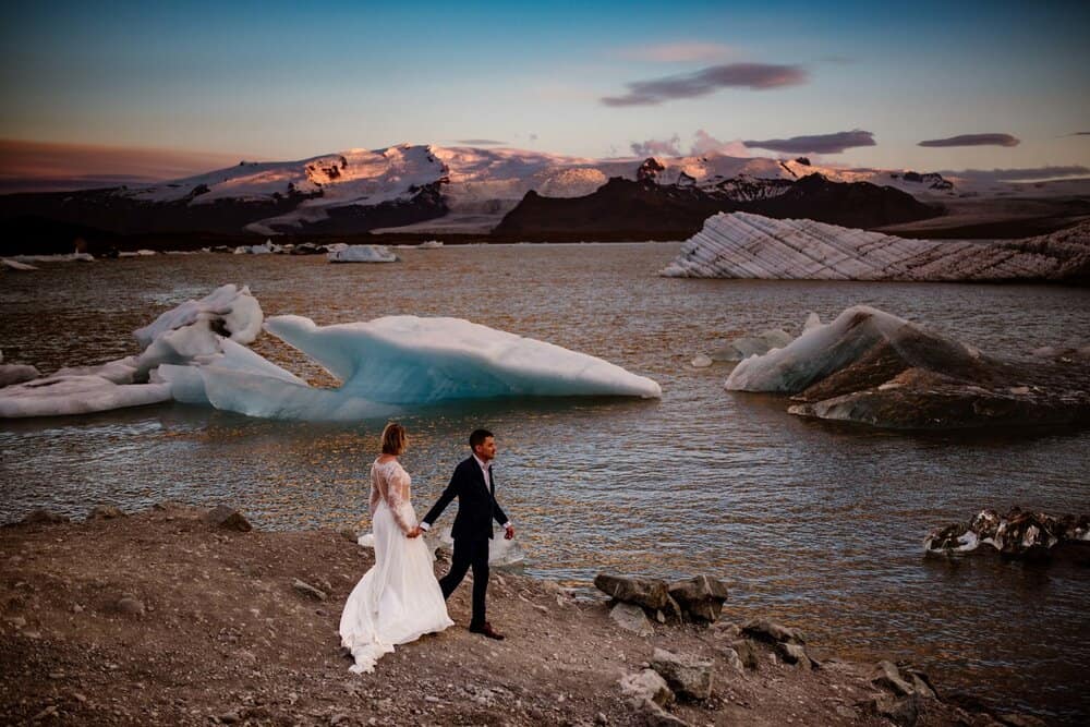 a bride and groom walking along the shoreline, looking at Iceland's water and glaciers.