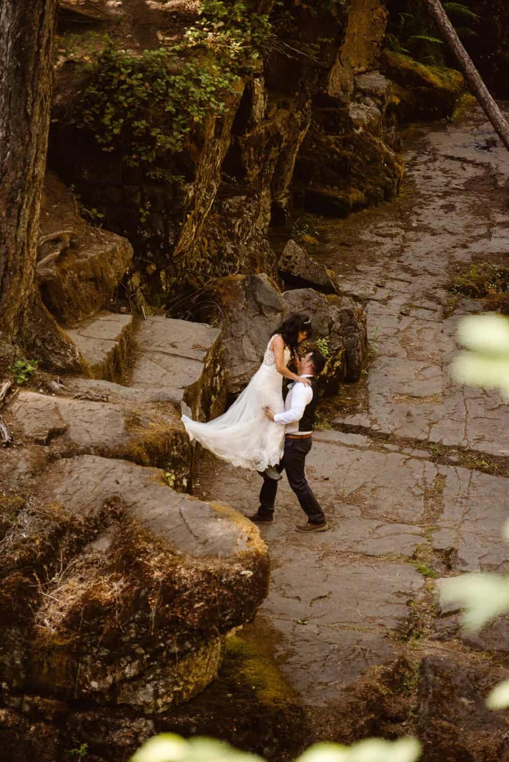 A groom helps a bride off of a rock ledge.