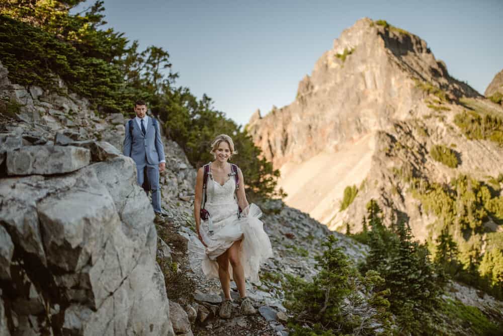 A bride smiles at the camera as she hikes in her wedding dress in the mountains.