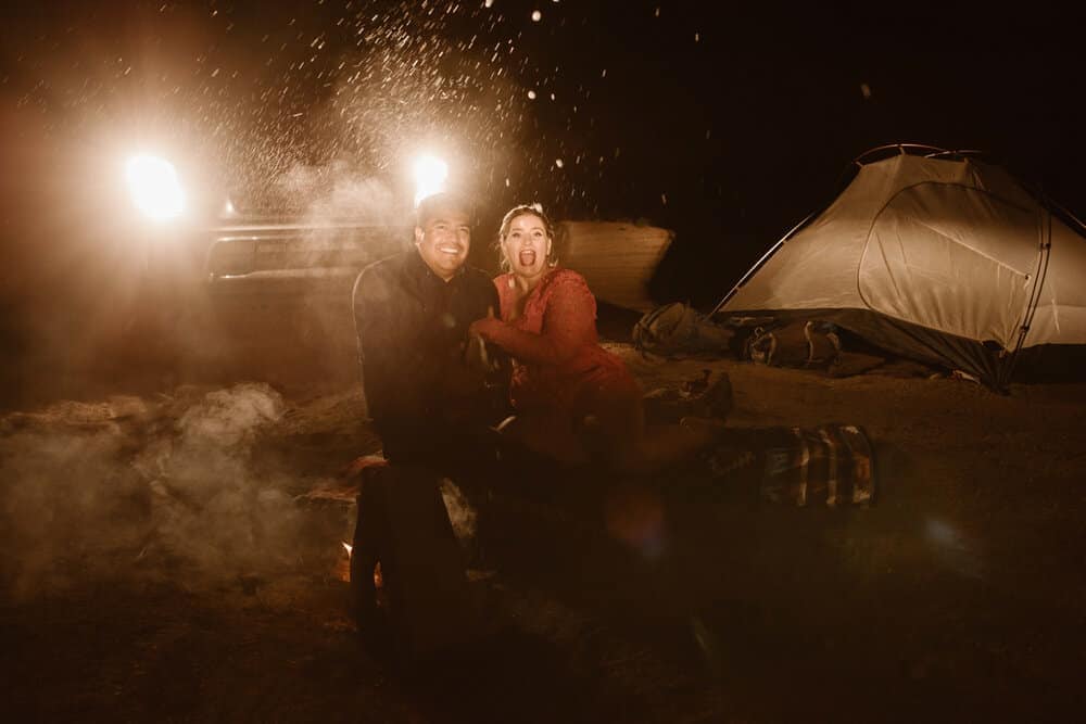 A couple at a camp fire together popping champagne to end the night.