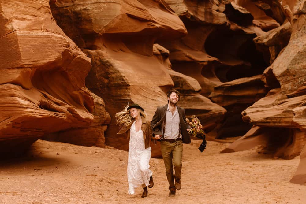 A couple runs through a slot canyon together holding hands and smiling. 