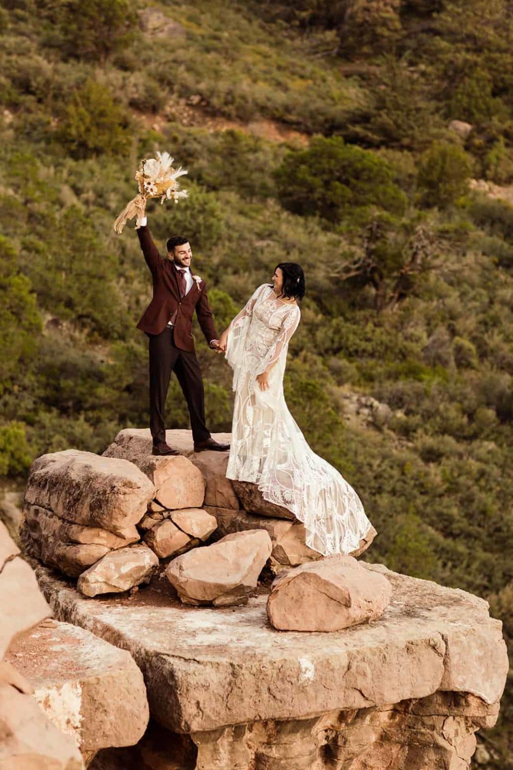 The groom holds up the brides bouquet in celebration as they stand on a limestone ledge