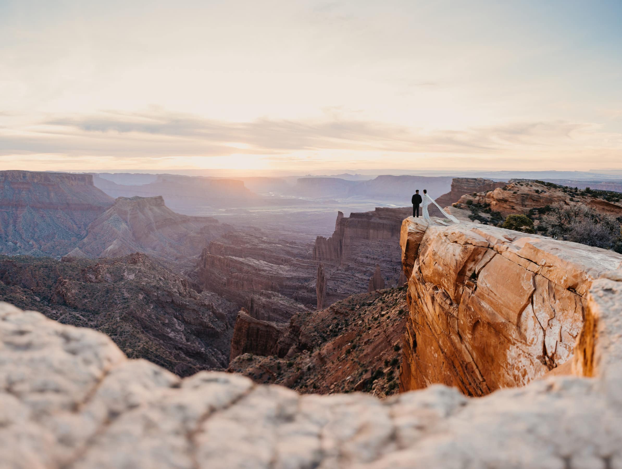 A couple stands at Top of the World off road route in Moab overlooking Fisher Towers at sunset.
