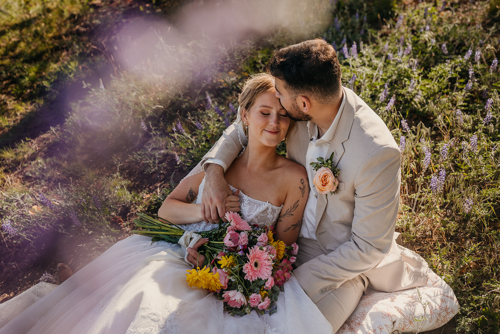 A groom holds his bride and kisses her forehead in a lupine field.
