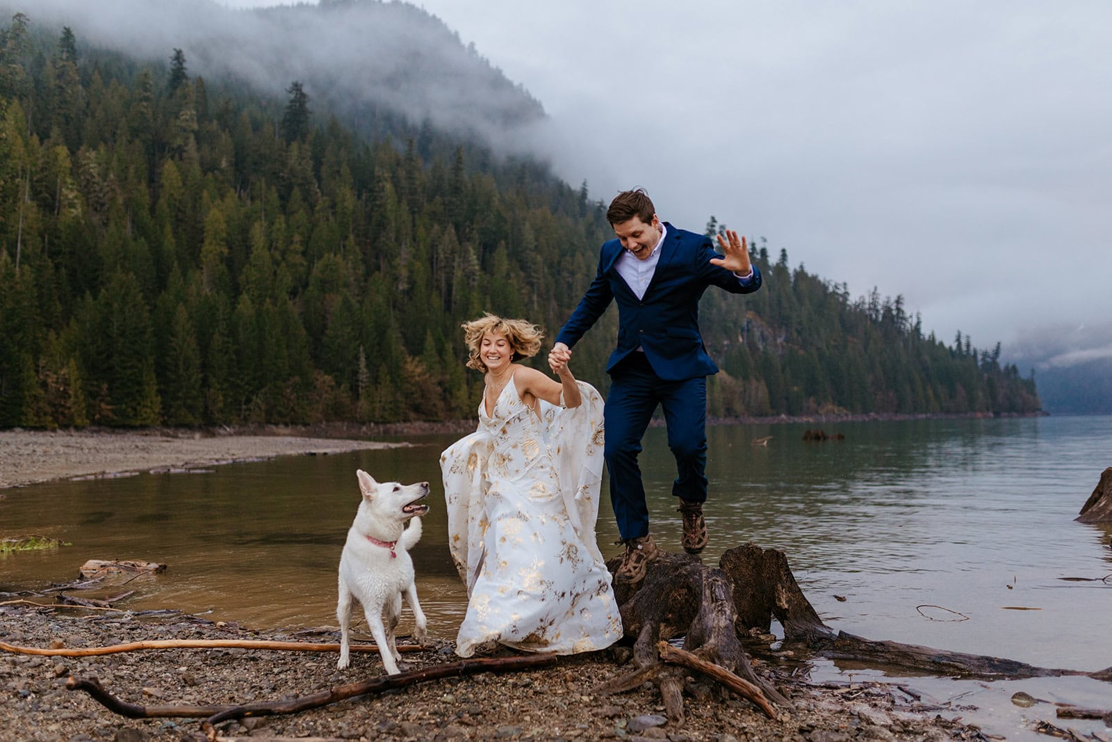 A couple jumps off a tree stump as their dog watches playfully.