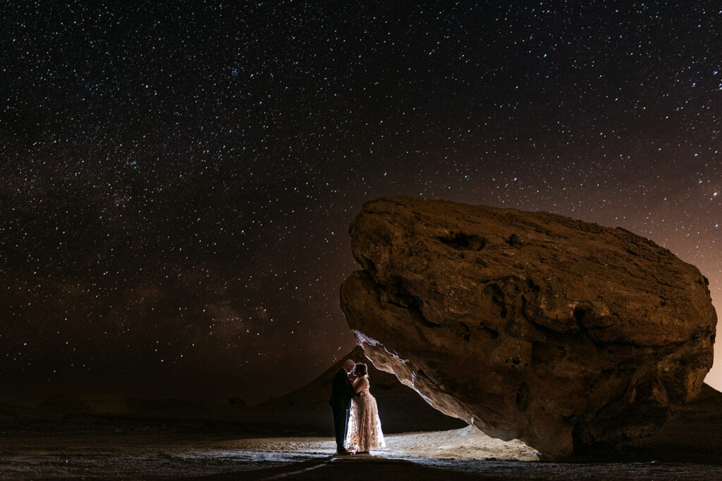 A couple stands under the stars together by a large boulder in Page, Arizona.