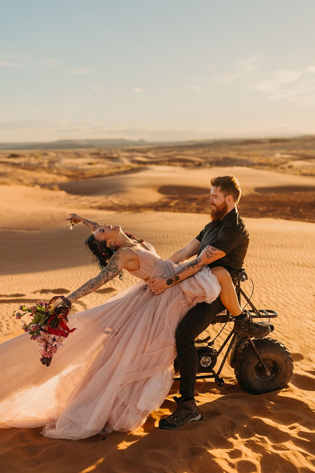 A bride leans back on a dirt bike as her groom holds her in the sand dunes.