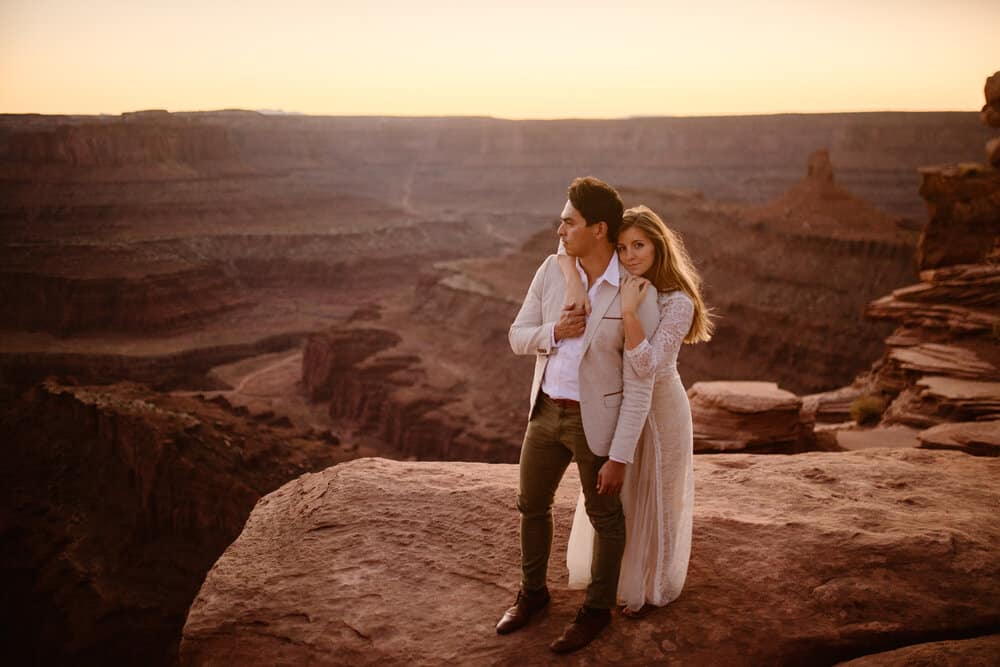 As a groom looks out at sunset the bride hugs him and smiles at the camera.
