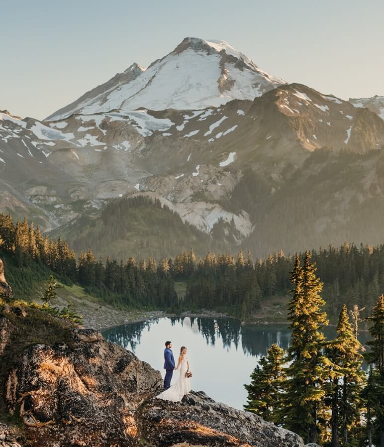 Two people on their elopement day hold hands while looking out at a view of a snow-capped mountain, trees, and a teal lake.