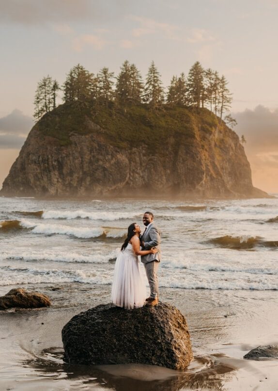 Two people hold one another on a rock with a beautiful view of an ocean and rock behind them. 