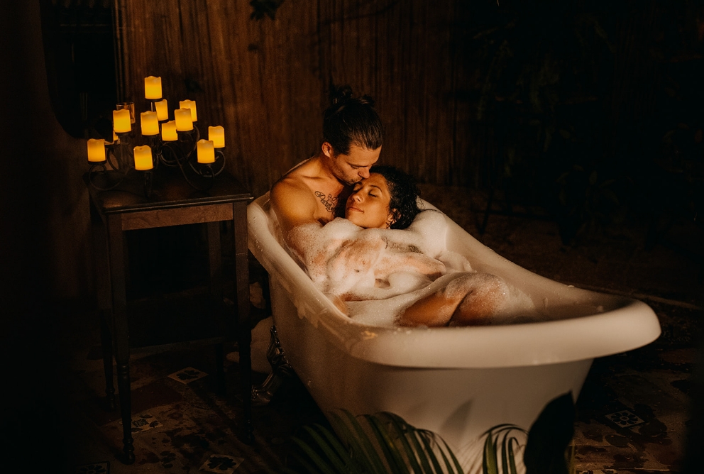 A couple sits in a bathtub with candles lit behind them.