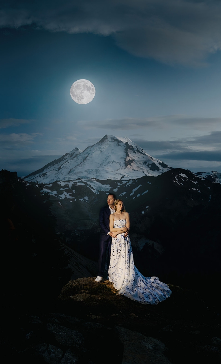 Two people hold one another under a clear sky with the moon shining brightly at Mt. Baker.