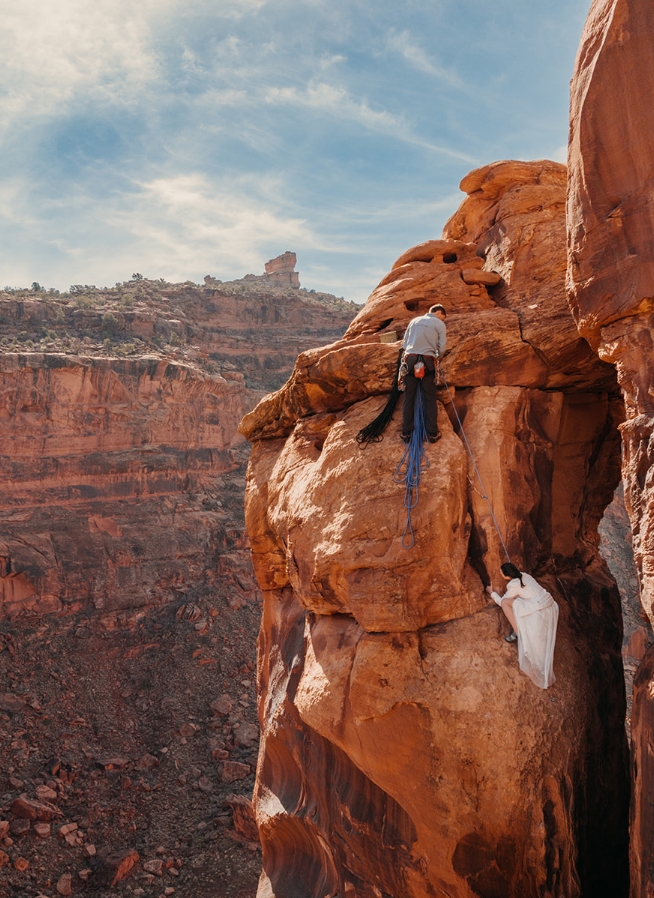 Two people in their wedding outfits climb a red rock.