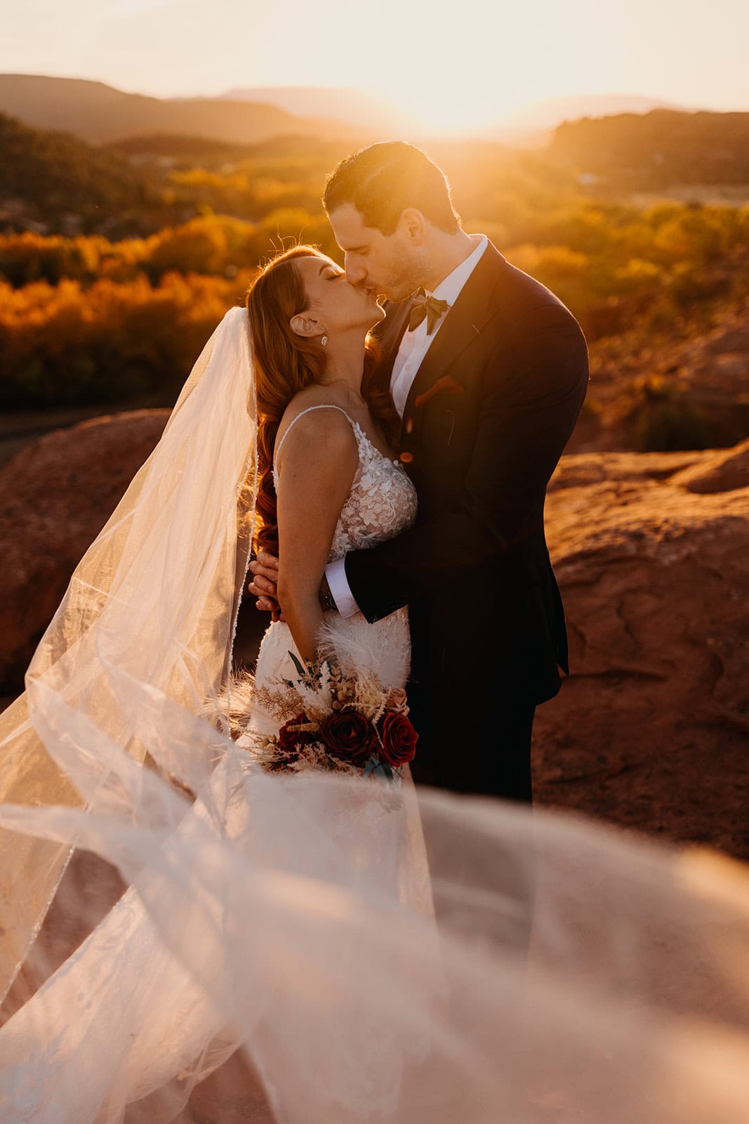 A bride and groom kiss at sunset as the bride's veil blows in the wind.