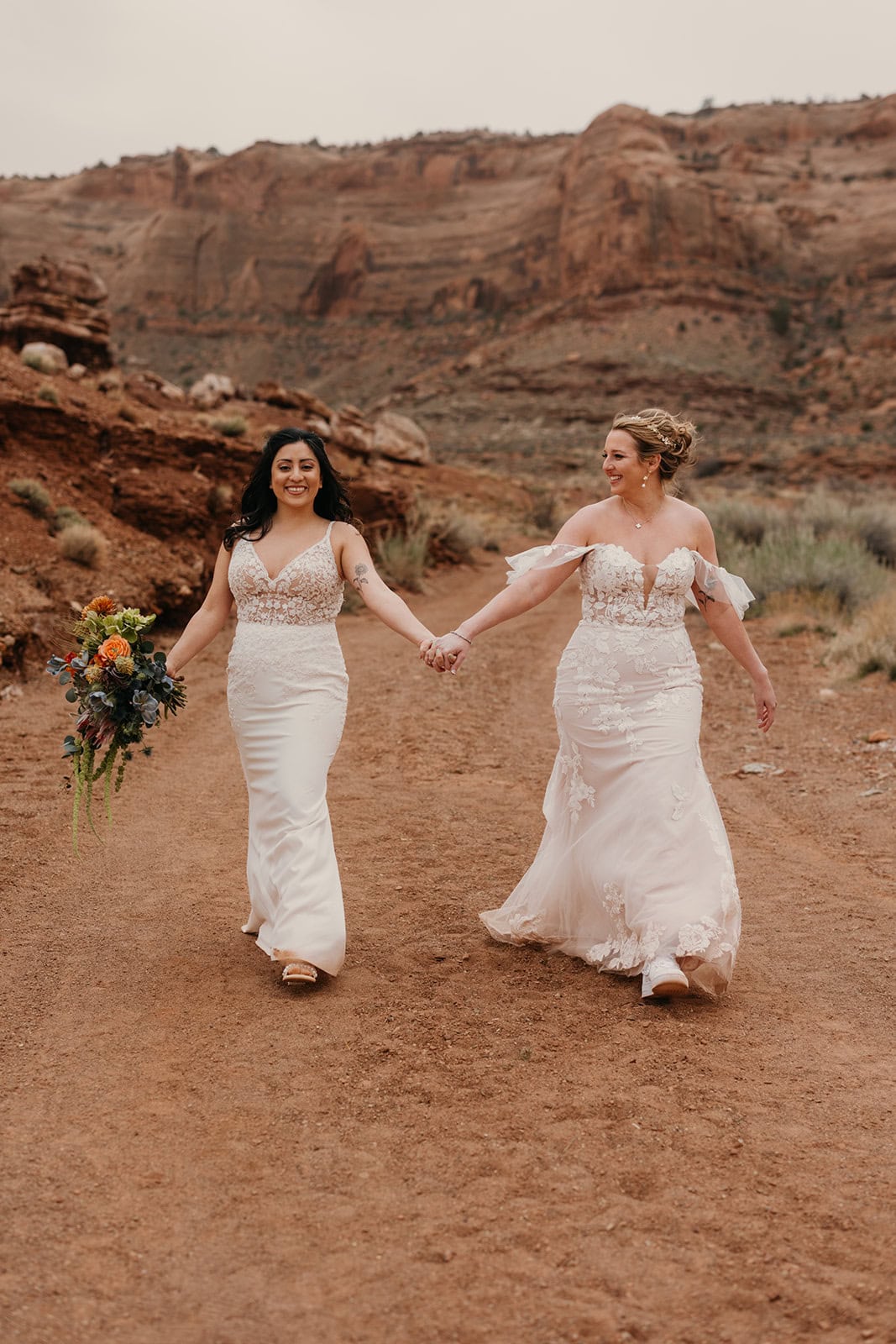 Two brides hold hands as they walk down a dirt road in Moab about to get married.