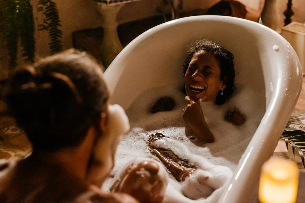 A woman laughs in a tub lit by candle light as her new husband makes a beard out of bubbles across from her.