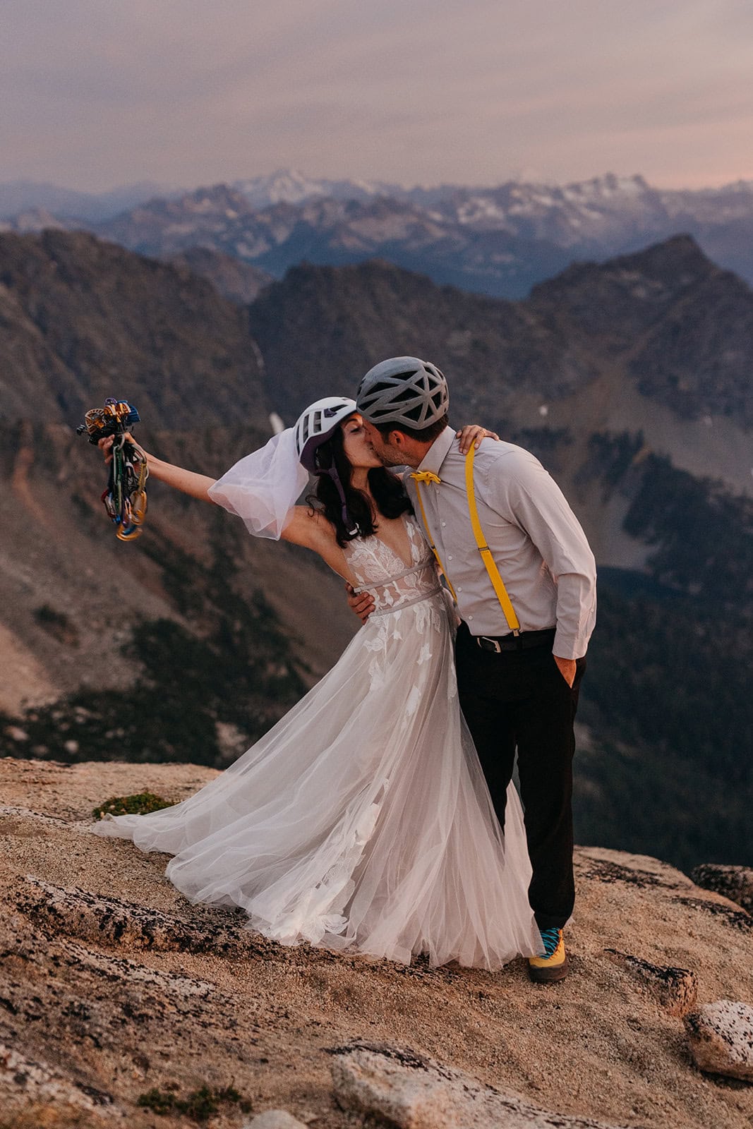 A bride fans her climbing cams out while her groom kisses her and they are both wearing helmets.