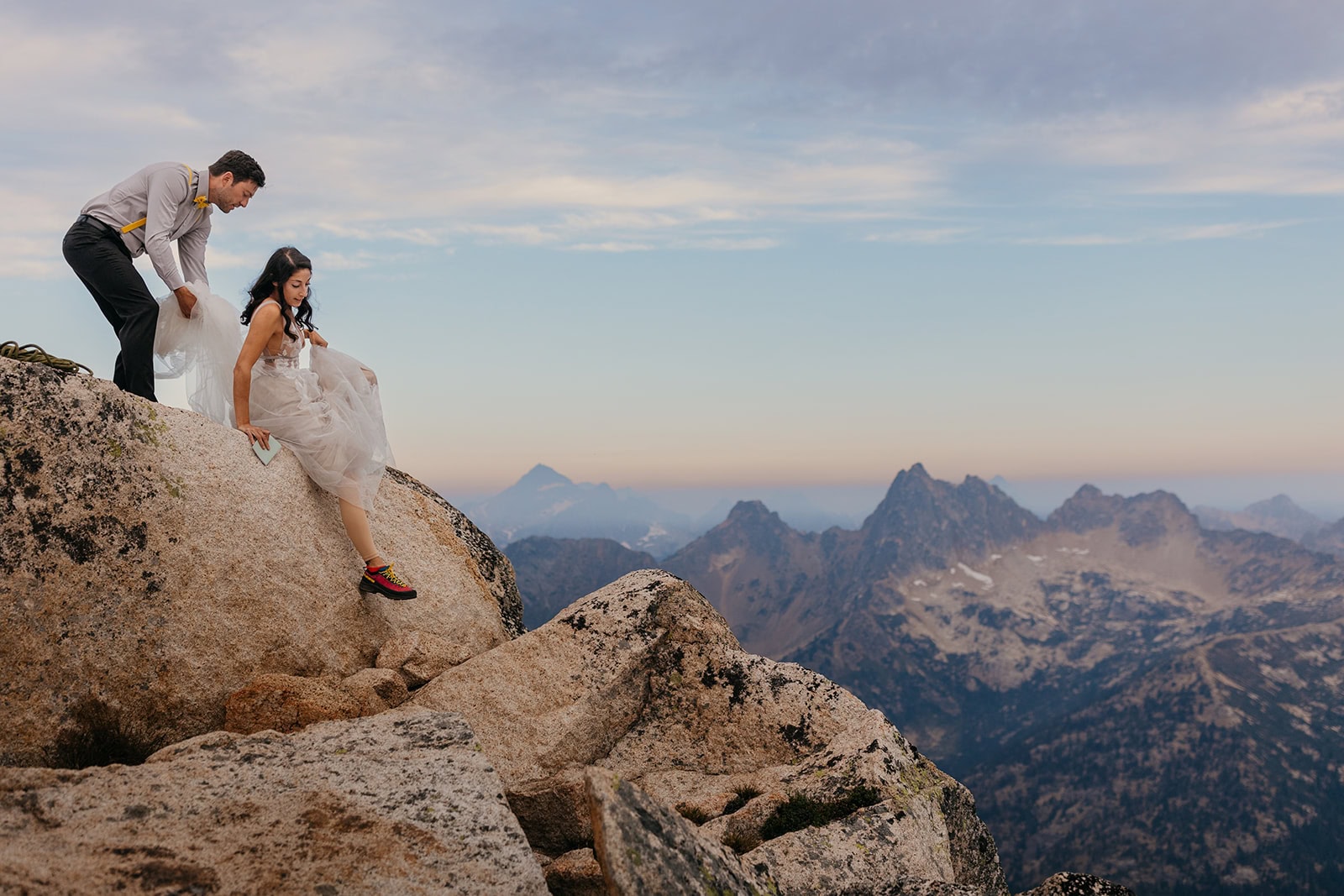 A bride and groom scramble across the summit of an alpine route in their wedding attire.