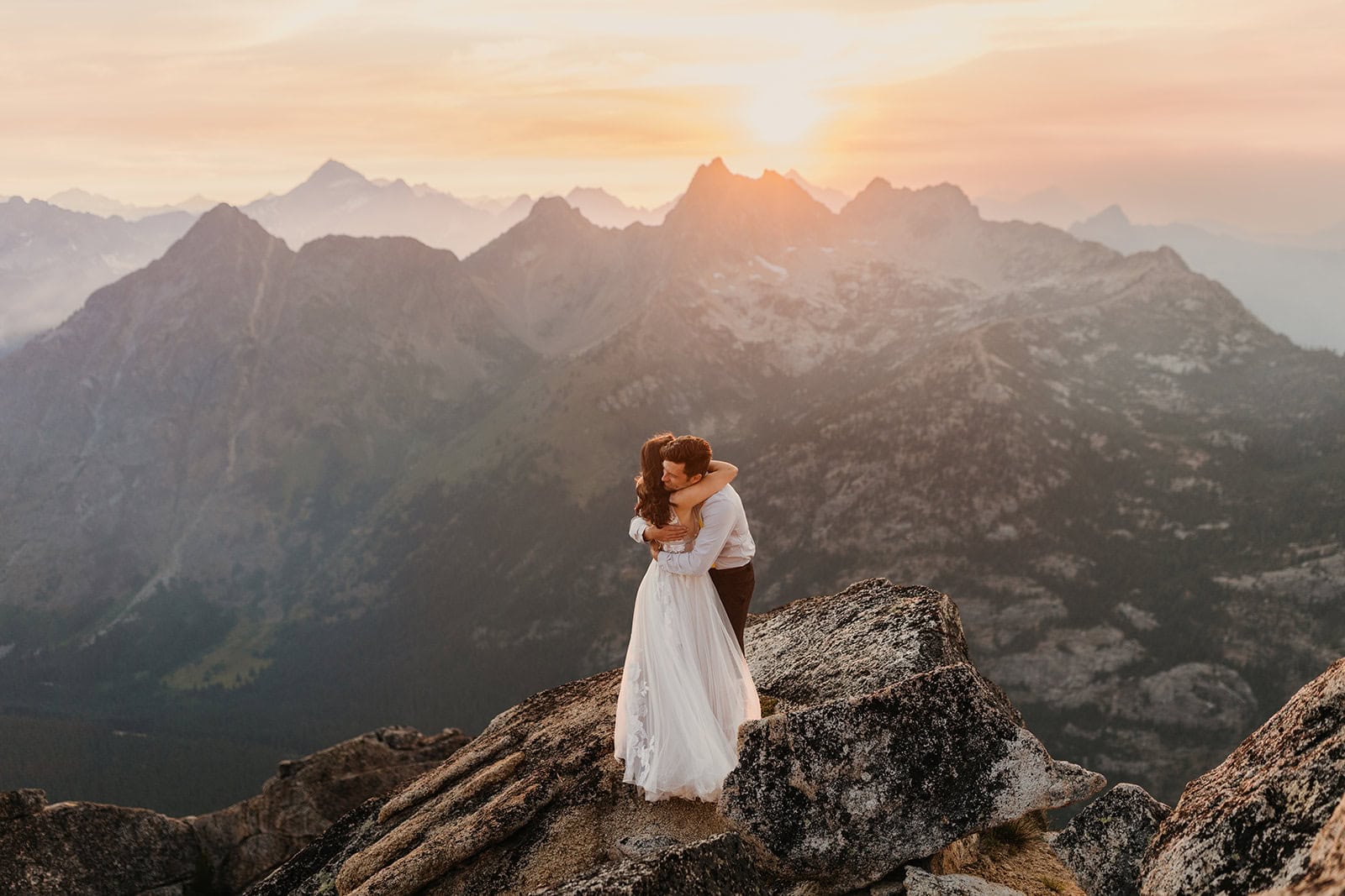 A bride and groom hug each other while the sun sets in the background