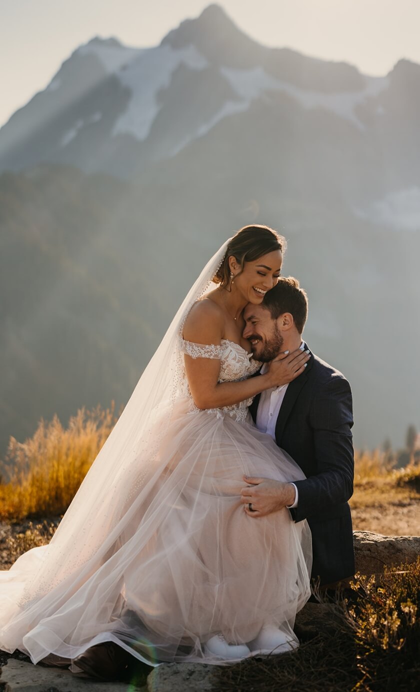 A person sitting on their partners lap as they laugh together on their wedding day in the mountains