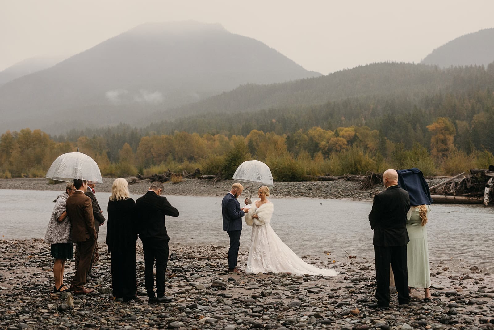 A couple shares their vows by a river with their family watching alongside them.