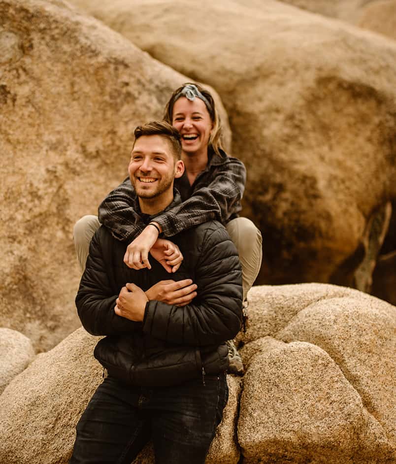 traci and bill laughing. traci has her eyes closed and is squatting behind bill on a rock.