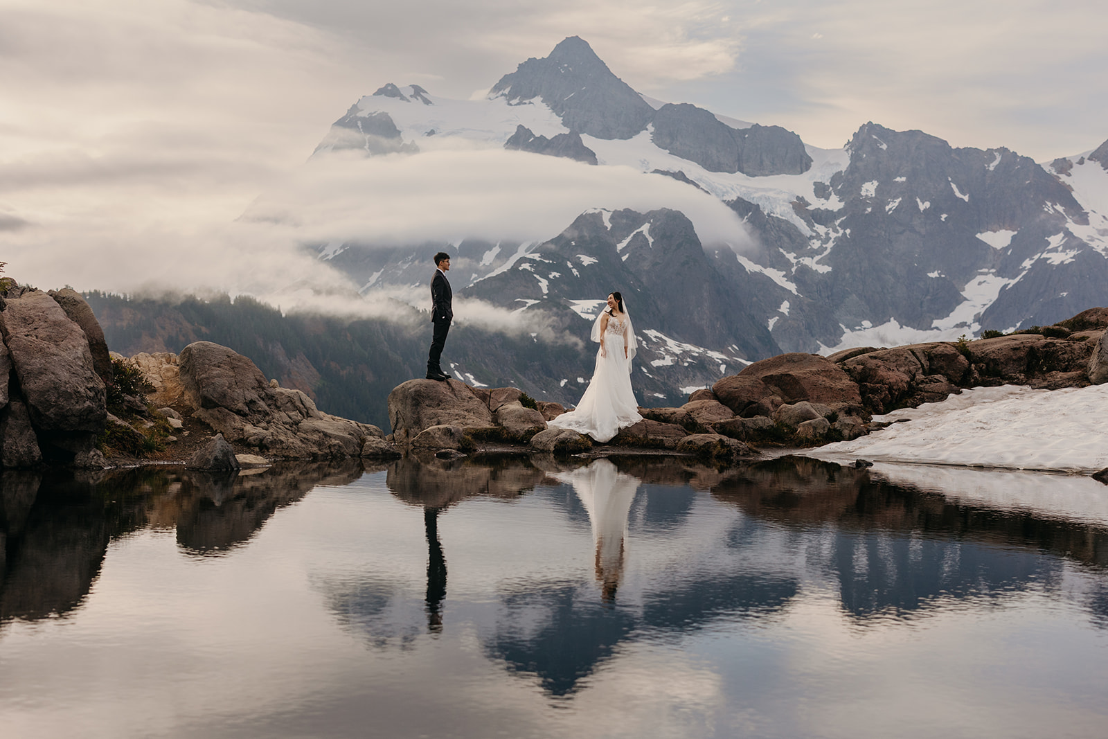 A couple pauses to look towards each other as they walk across a snow melt tarn in Mt Baker.