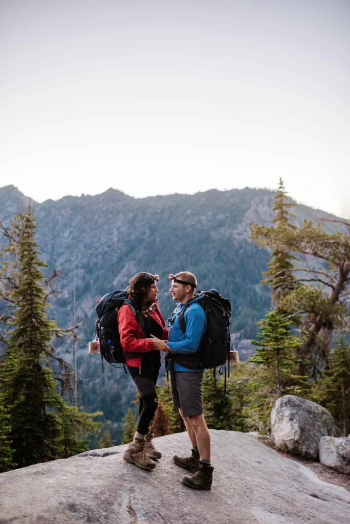 A couple stands together as the sun starts to set and they hike down the mountain.